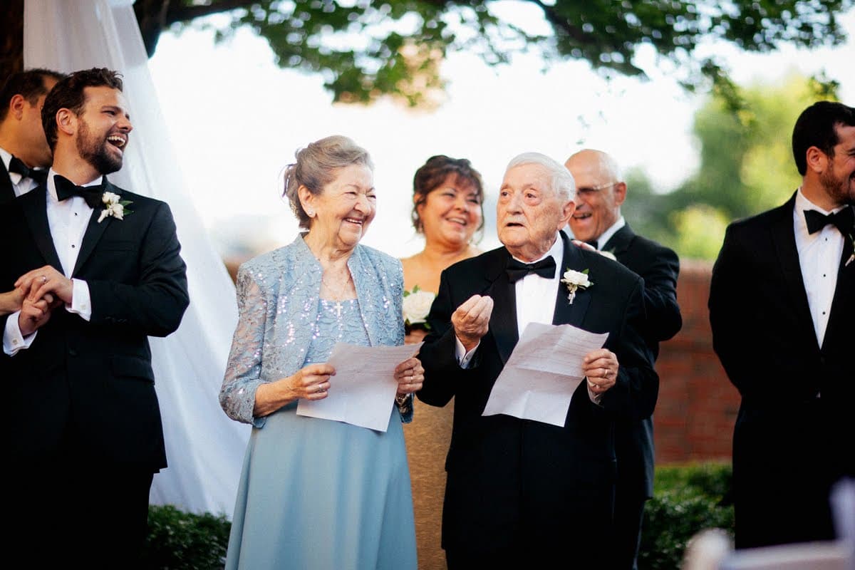 Pino Cirincione laughing with his wife at a family wedding, both holding papers, surrounded by family in formal wear